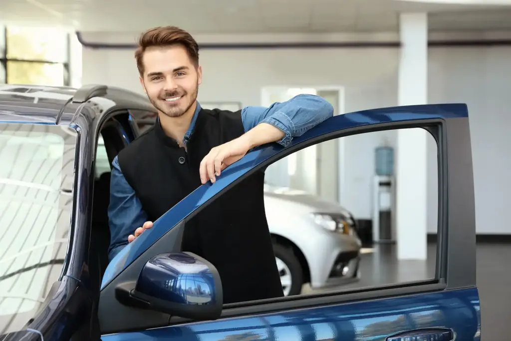 junger Mitarbeiter im Autohaus lehnt an geöffneter Autotür und lächelt in die Kamera - Stellenangebot Kfz-Serviceassistent
