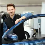 junger Mitarbeiter im Autohaus lehnt an geöffneter Autotür und lächelt in die Kamera - Stellenangebot Kfz-Serviceassistent
