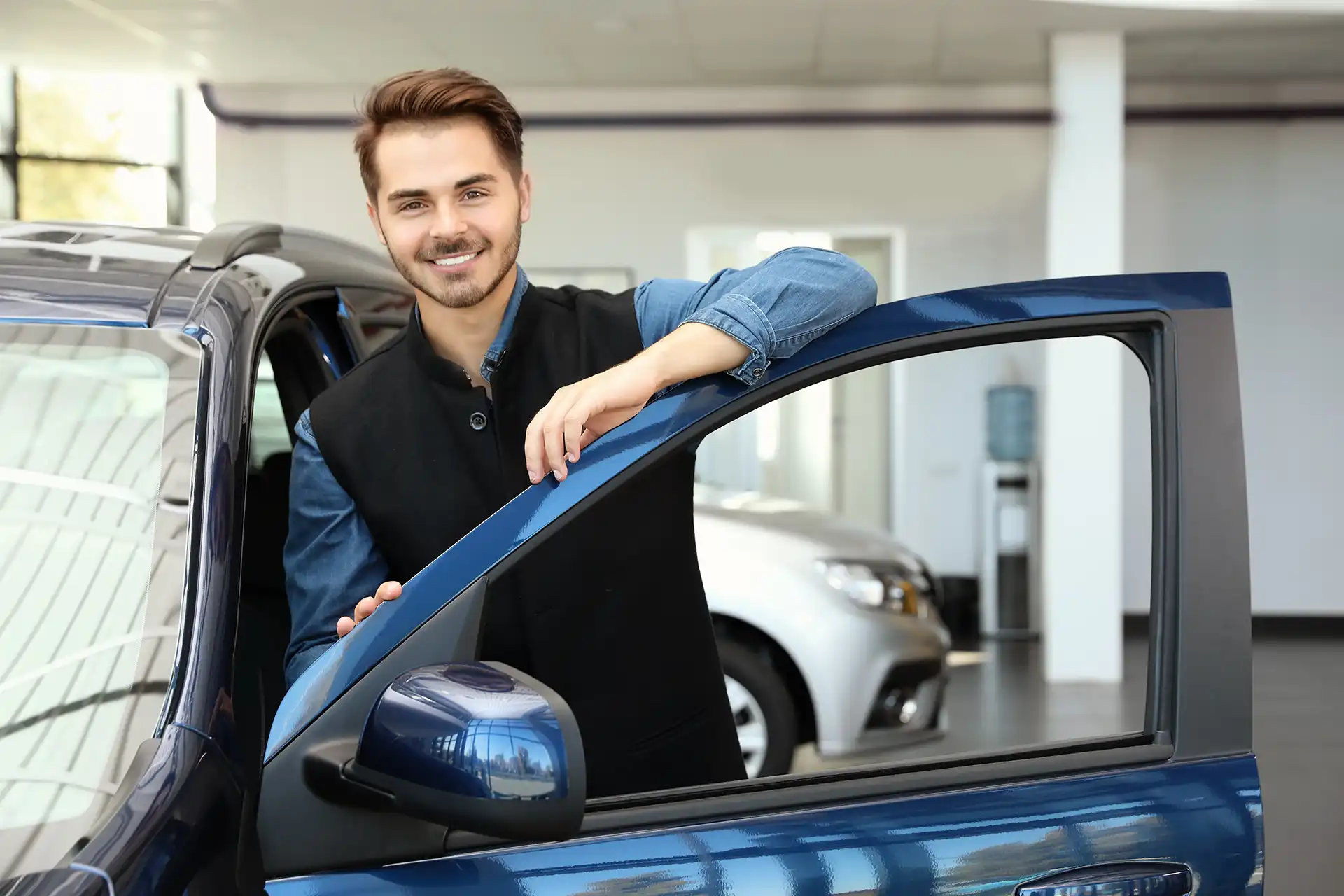 junger Mitarbeiter im Autohaus lehnt an geöffneter Autotür und lächelt in die Kamera - Stellenangebot Kfz-Serviceassistent
