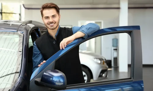 junger Mitarbeiter im Autohaus lehnt an geöffneter Autotür und lächelt in die Kamera - Stellenangebot Kfz-Serviceassistent